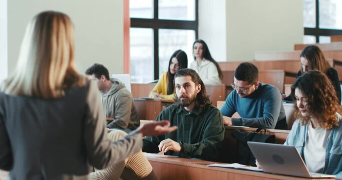 Back view on female professor at University lection talking and explaining material to multiethnic students in classroom. Woman teacher having speech. Guy student asking question. Rear.