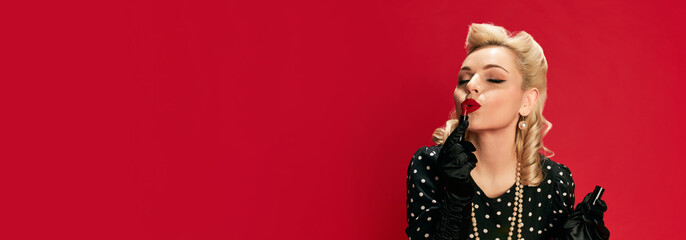 Beautiful young girl with stylish hairstyle posing in dress, applying red lipstick against red...