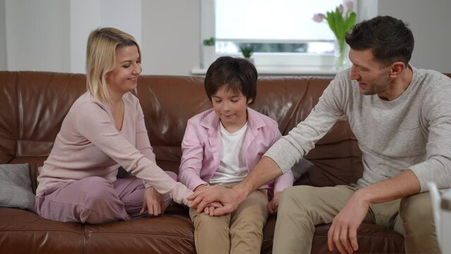 Supporting parents stacking hands with upset son gesturing thumbs up smiling. Confident Caucasian man and woman endorsing sad boy sitting on couch in living room at home