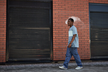 Side view of young black man in casualwear walking along brick wall of building and blowing cloud of vapor out of his mouth