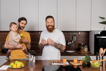 Cheerful gay man holding cup near partner and daughter with apple in kitchen.