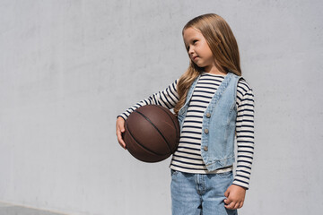 preteen girl in denim vest and blue jeans holding basketball near mall building.