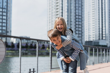 happy preteen boy in denim outfit piggybacking girl on river embankment.