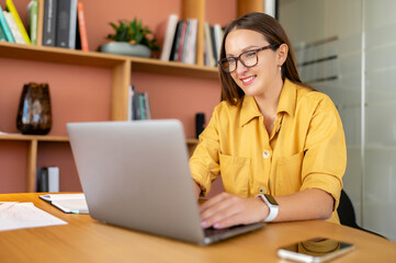 Fototapeta na wymiar Portrait of young woman in glasses sitting at the desk with laptop, typing an email, messaging colleague, chatting, discussing work moments with pleasant smile