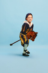 Happy cute little boy dressed up as medieval little prince and pageboy ride toy horse over light...