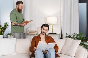 Gay man reading book near partner in living room at home.