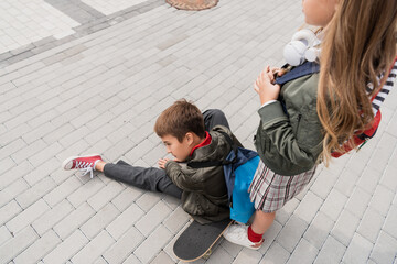 overhead view of preteen boy in stylish bomber jacket sitting on penny board near girl in wireless...