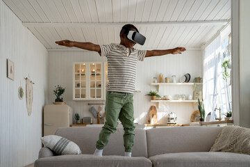 Carefree smiling African American child boy play video game with virtual reality goggles pretending...
