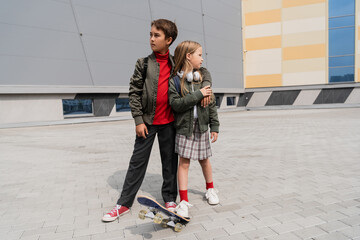 full length of preteen boy in stylish bomber jacket hugging girl in skirt while standing near penny...