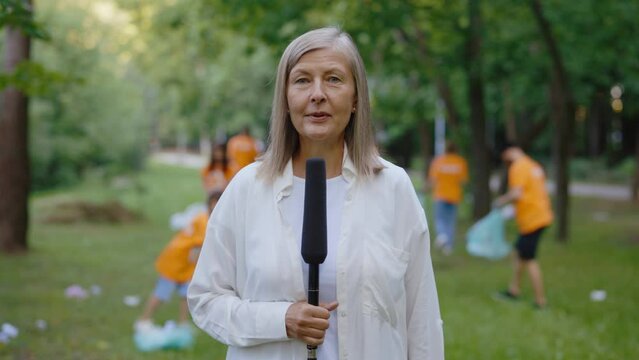 Female Journalist Reports on How Volunteers Collect Garbage and Clean Up the Planet. News. Group of Eco Activists Volunteers Cleaning Park from Trash in the Park on Background