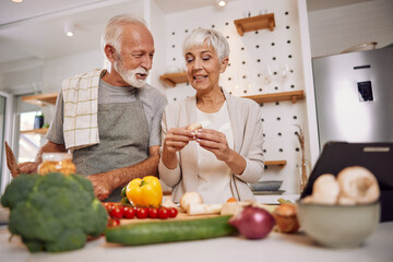Smiling elderly vegan couple preparing a fresh vegetable salad in the kitchen, using a tablet