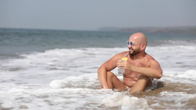 Video positive relaxed bald bearded man in sunglasses and shorts drink cocktail with straw and lemon, sit in sea waves