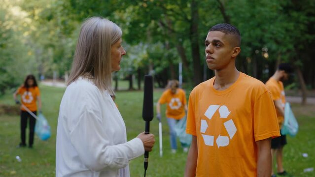 Female News Reporter Standing in Polluted Forest and Interviewing Eco Volunteer Who Are Collecting Rubbish. News. Group of Eco Activists Cleaning Park from Trash in the Park on Background