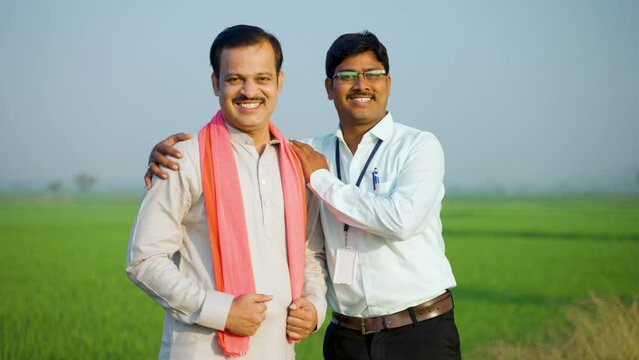 Happy supportive banker with village farmer standing by looking at camera near green farmland - concept of banking service, assistance and assurance