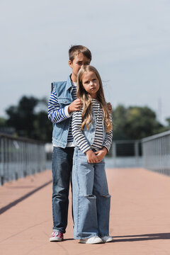 full length of preteen boy and girl in denim clothes standing on riverside embankment.