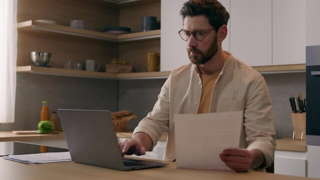 Caucasian man husband male homeowner householder renter guy businessman with papers pay bills online on website app paperwork planning budget check finances mortgage payments using laptop at kitchen
