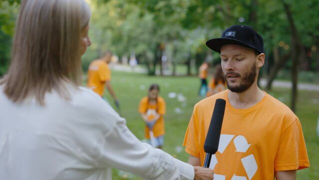 Female News Reporter Standing in Polluted Forest and Interviewing Caucasian Eco Volunteer Who Are Collecting Rubbish. News. Group of Eco Activists Cleaning Park from Trash in the Park on Background.