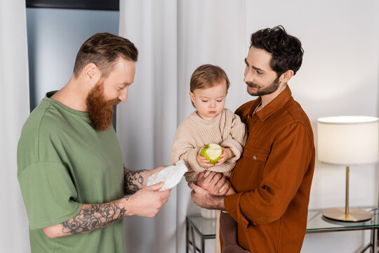Positive gay couple holding baby daughter and diaper at home.