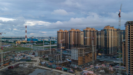 Naklejka premium Aerial view of a construct residential high-rise buildings, cranes, steel, concrete, cargo, work next to park and highway in the St. Petersburg at clouds summer afternoon, shadows