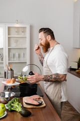 Side view of tattooed man in apron tasting food while cooking in kitchen.
