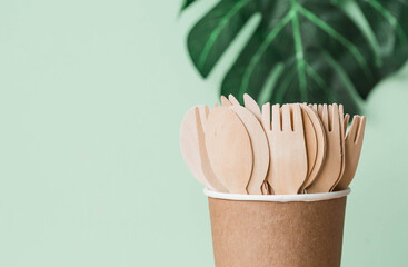 Eco-friendly disposable kitchen utensils in a paper cup on a beige-green background. Wooden forks, spoons and knives. Ecology, the concept of zero waste. - obrazy, fototapety, plakaty