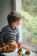 Child playing with colorful toy blocks. Educational toys for young children and looking on the rain drops on the window