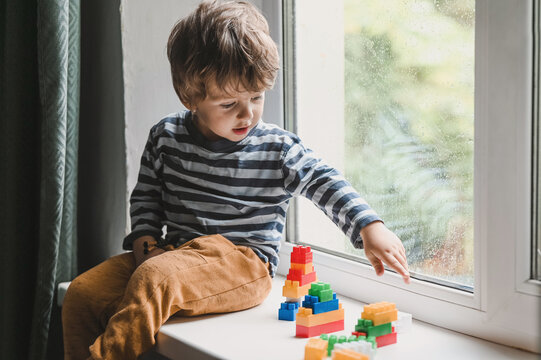 Little boy sitting on the window sill and  playing with lots of colorful plastic blocks constructor. Boy playing with construction blocks at kindergarten. looking on the rain drops on the window 