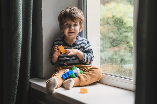 Little boy sitting on the window sill and  playing with lots of colorful plastic blocks constructor. Boy playing with construction blocks at kindergarten. looking on the rain drops on the window