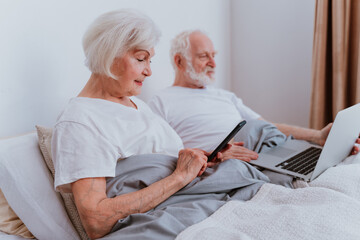 Obraz premium Elderly couple lying in bed at home and using computer laptop and smartphone