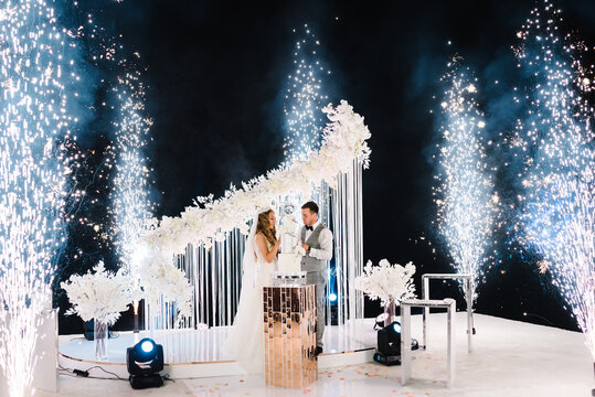 Bride and a groom is cutting and eating wedding cake. Hands cut the cake with flowers. Newlywed near arch. Night ceremony with light and fireworks. Fire show.