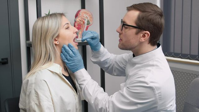 Young male ENT otorhinolaryngologist doctor in white coat holding tongue depressor examing female patioen throat during visit in privat clinic