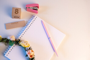 Material de oficina rosa con una corona de flores descansando sobre un cuaderno sobre un fondo rosa para el Día Internacional de la Mujer. - obrazy, fototapety, plakaty