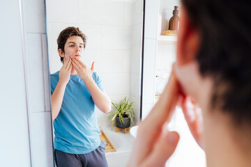 Concentrated caucasian teenage boy with acne problem at his face looking in mirror at home bathroom. Teenager skin care every day treatment process. Selective focus