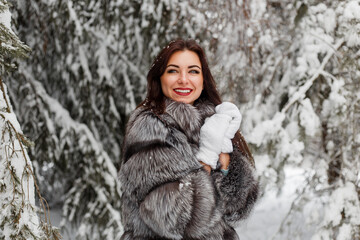 A happy smiling young lady in a fur coat and knitted mittens stands in a snowy forest. Joyful winter mood