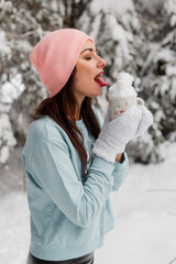 A young lady in a sweater, hat and knitted mittens in a snowy forest holds a mug of snow in her hands and licks it off. Cheerful winter mood