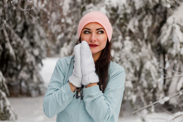 A young happy lady in a sweater, hat and knitted mittens stands in a snowy forest among the fir trees. Fairytale and joyful winter mood