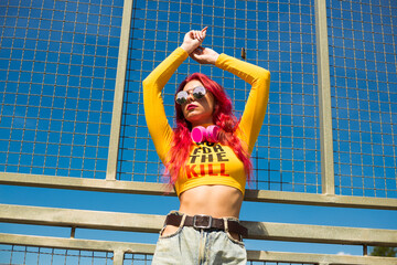 A lady with pink hair, headphones around her neck, in a yellow top, ripped jeans and sunglasses is standing on the street with her hands up. Vintage retro clothing style of the 2000s in modern fashion