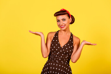 A happy smiling lady in a polka dot dress with a retro hairstyle and red lipstick stands on a yellow background. A pin-up look with a modern twist. Vintage party