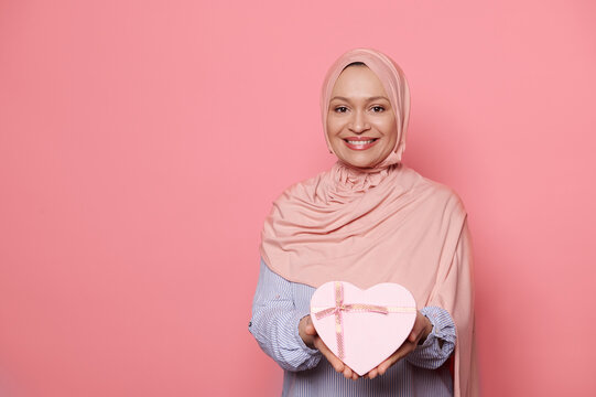 Middle-aged Muslim Woman Dressed In Elegant Pink Hijab And Blue Casual Shirt, Smiling A Beautiful Toothy Smile, Holding Out At Camera A Cute Heart Shaped Pink Gift Box, Isolated Bright Pink Background