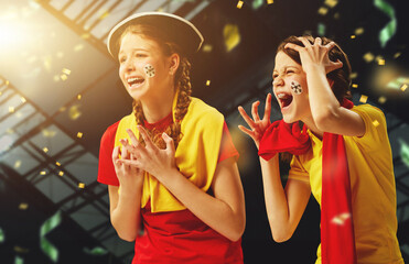 Tense game moment. Young girls emotionally watching football match, cheering team over dark background with confetti. Concept of sport, leisure time, emotions, hobby and entertainment