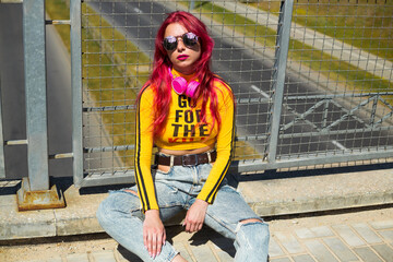 A lady with pink hair in sunglasses, a yellow top and ripped jeans sits on the sidewalk. Vintage retro style clothing from the 2000s in modern fashion. The return of old trends in a new way
