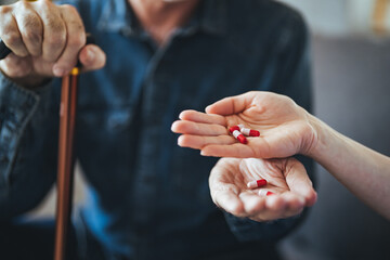 A home healthcare worker helping a senior man in his 70s take his prescription medicine. The patient is in the kitchen, holding a cup of water, looking down as the nurse puts the pills in his hand.