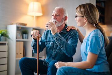 A young nurse sits at a table in the living room with a senior man and provides him with medical assistance. Female caregiver giving the medicine to her older male patient