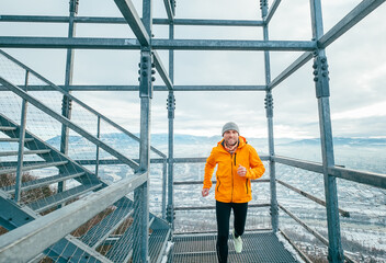 Smiling runner man dressed in bright orange softshell sporty clothes running down by huge steel industrial stairs in cold winter day. People healthy lifestyle concept photo.