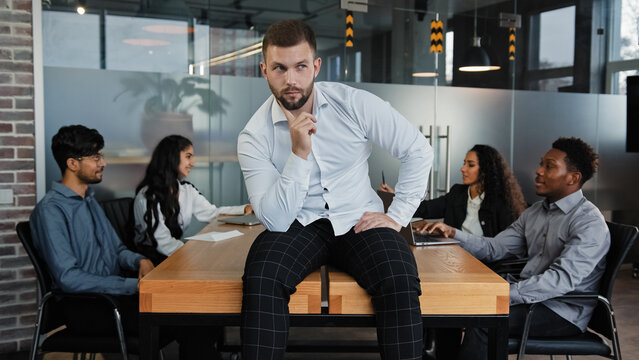 Thoughtful thinking pensive man leader boss businessman think about startup idea company CEO sitting on office table on background of multiracial business team workers group discuss project teamwork