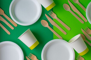 Eco-friendly disposable dishes made of bamboo wood and paper on a green background. Draped spoons,...
