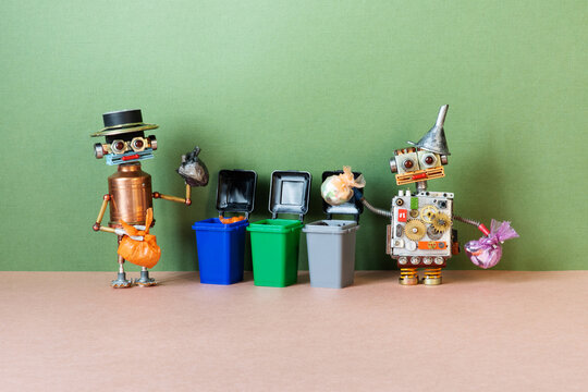 Two toy robots with garbage bags pose near street trash cans, dumpsters. The concept of separate storage of garbage in specialized containers of different colors.