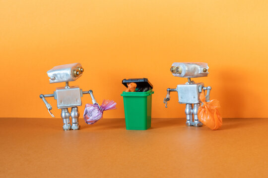 Two small silver metal robots are holding garbage bags ready to be disposed of in a dumpster, trash can. Orange background.