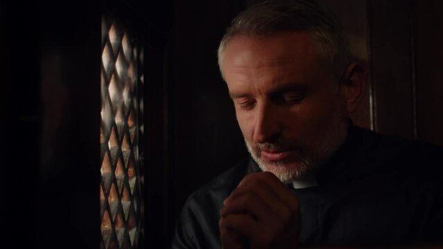 The black-robed priest folded his hands in prayer. The gray-haired pastor prays while sitting in the confessional.