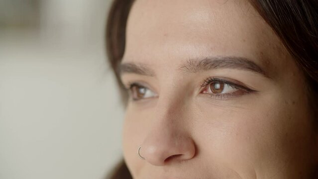 Cropped view of female face with brown eyes. A brunette woman with a nose piercing raises her eyes and looks away.